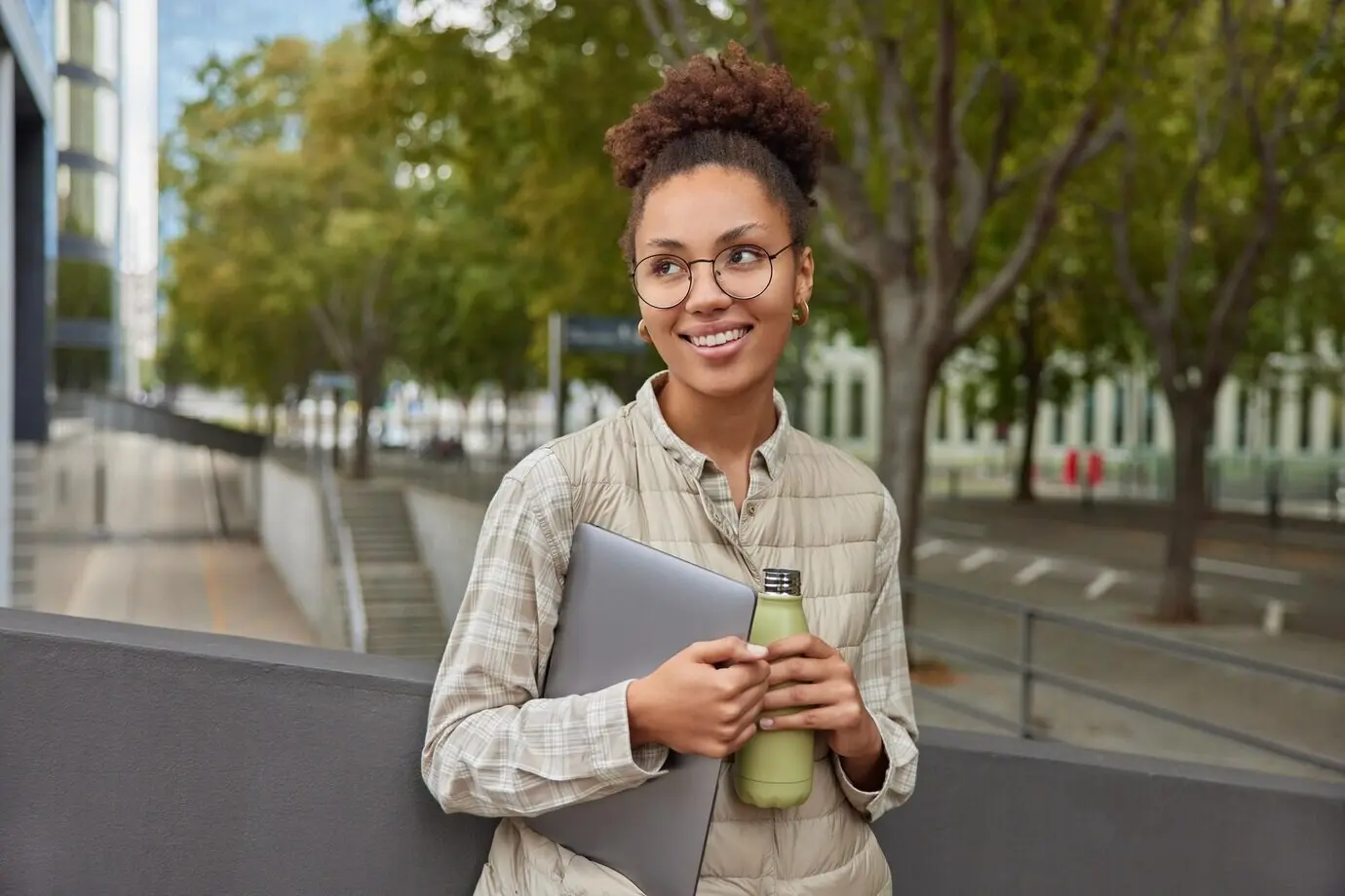 Außenaufnahme einer nachdenklichen, gut gelaunten jungen Frau, die sich umschaut, während sie durch die Stadt geht und dabei einen Tablet-Computer sowie eine Flasche frischen Wassers trägt.